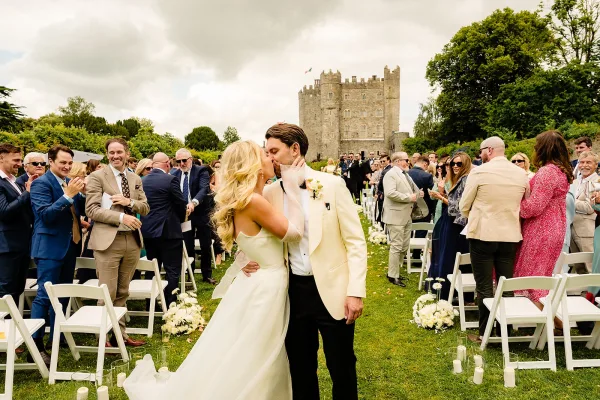 Ceremony kiss as newlyweds walk the outdoor wedding ceremony aisle, guests cheering between flower-lined rows with a stone castle behind