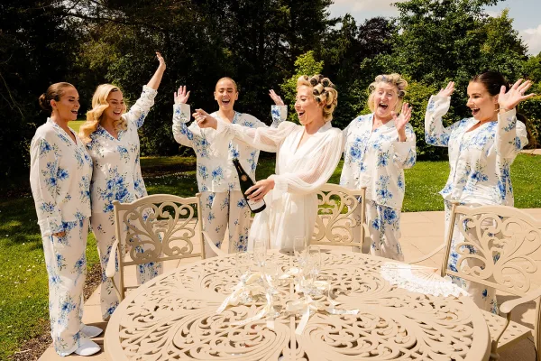 Bride getting ready with bridesmaids getting ready in matching pajamas, popping champagne on a stone patio with garden trees in daylight