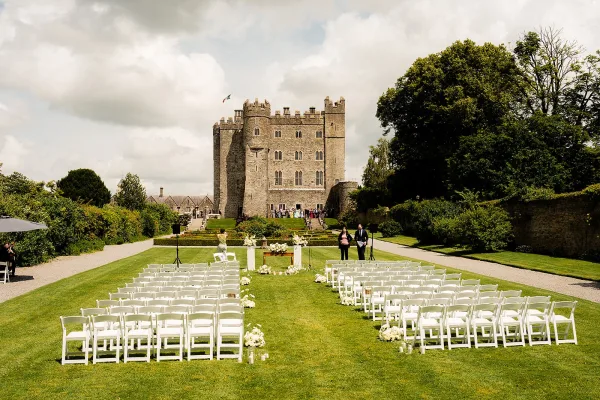 Outdoor ceremony setup with garden wedding ceremony seating, white folding chairs and floral-lined aisle leading to a castle backdrop