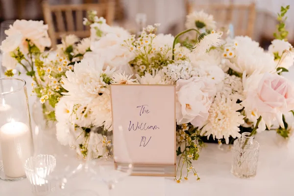 Wedding table centerpiece with a white floral centerpiece of daisies and greenery, beside floating candle cylinders and an acrylic table number sign at an indoor reception table