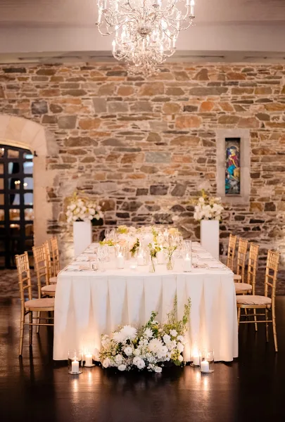 Reception tablescape with white tablecloth, low floral centerpiece, and candlelit glass votives beneath a chandelier in a stone-walled hall