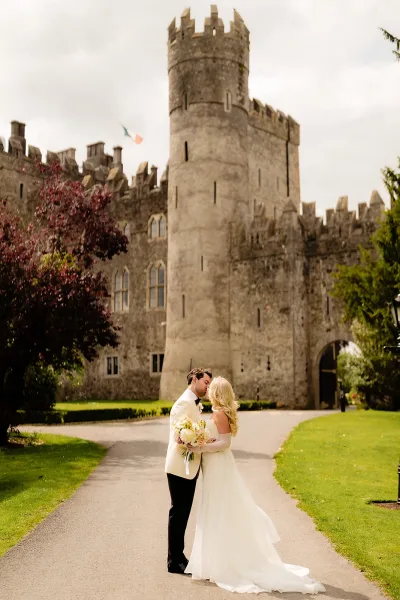 Wedding kiss portrait of bride and groom kissing, holding a bouquet in front of a stone castle archway under cloudy sky