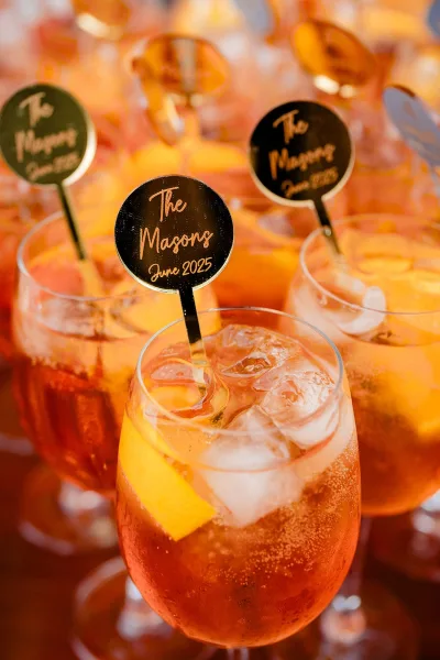 Wedding signature drinks with custom cocktail stirrers and orange peel garnish in stemless glasses, set against a softly blurred bar setup