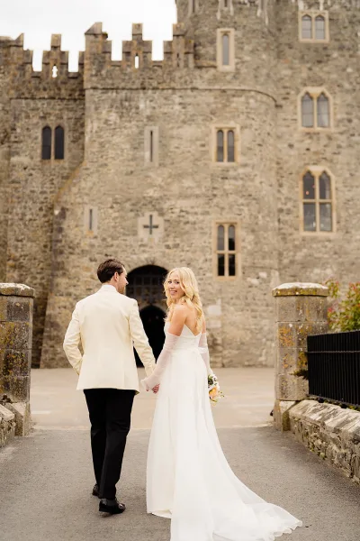 Couple portrait of bride and groom walking away hand in hand, bride looking back with veil and long train by a stone castle archway