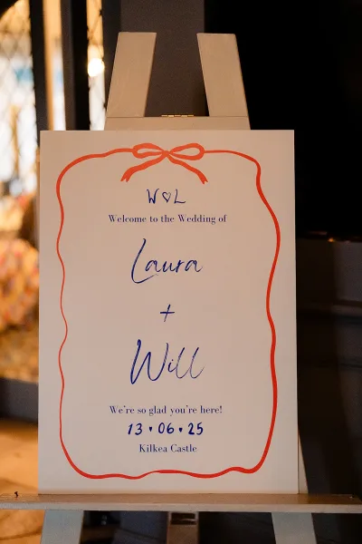 Wedding welcome sign with calligraphy welcome sign text on an easel, ribbon border graphic, lit by window light against a dark wall
