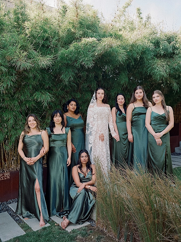 Bridesmaid group portrait with bride and bridesmaids in emerald green satin dresses, bride in lace gown and veil before bamboo hedge
