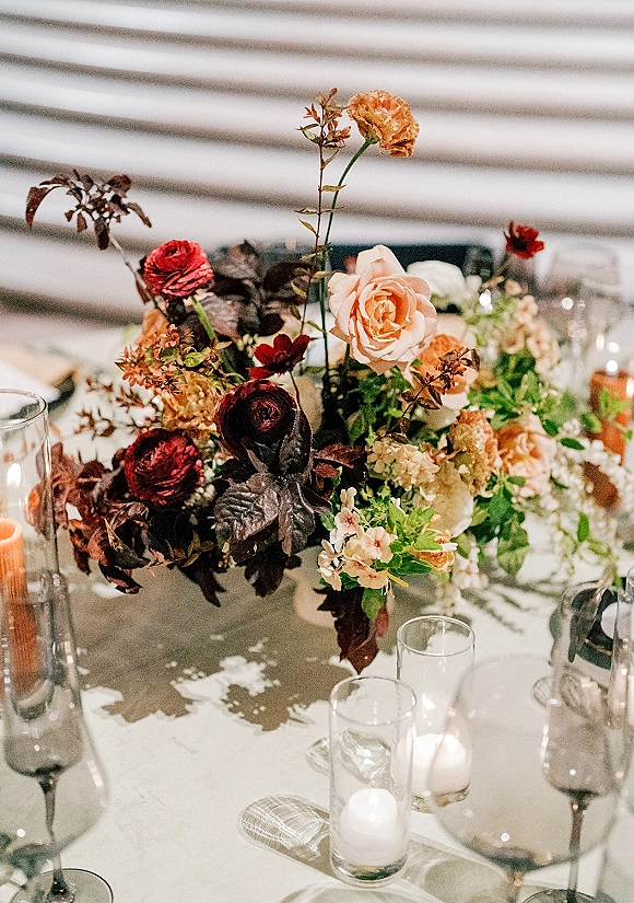 Wedding centerpiece with lush floral arrangement of roses and ranunculus, dark foliage, and floating candles in glass vases on a reception table