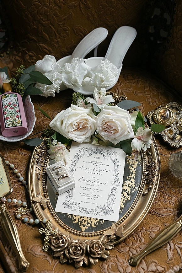 Invitation suite wedding invitation flat lay with ornate gold frame, pearl heels, rings, white roses, jewelry and perfume on bronze brocade fabric