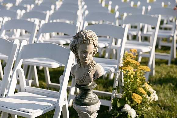 Ceremony aisle decor with outdoor wedding ceremony chairs, low white and yellow florals with greenery lining a grass lawn beside a stone bust statue