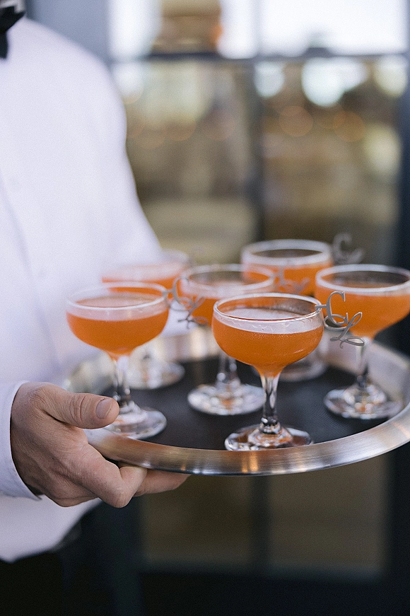 Wedding cocktails served as a signature wedding cocktail in coupe glasses, orange drinks on a metal tray with garnish picks at a blurred indoor bar
