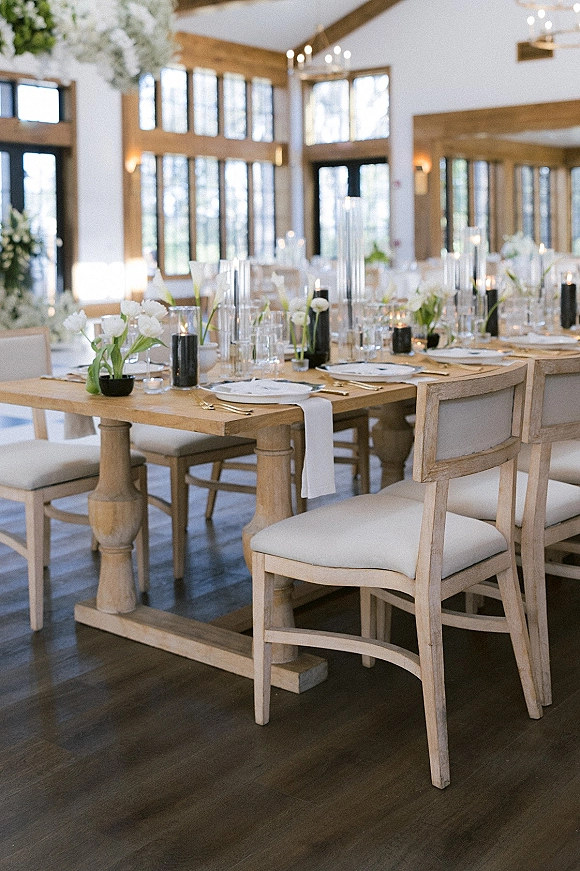 Reception tablescape on a farmhouse wedding tablescape with white runner, taper candles, glass vases, gold flatware, and window-lit room