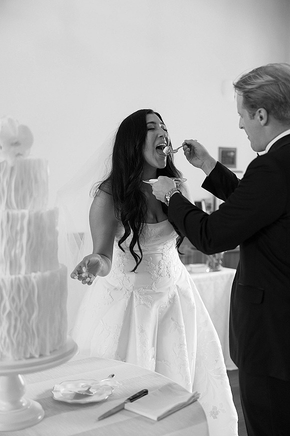 Wedding cake cutting as bride and groom slice a tiered white cake with bow topper at an indoor reception dessert table, cake knife in hand