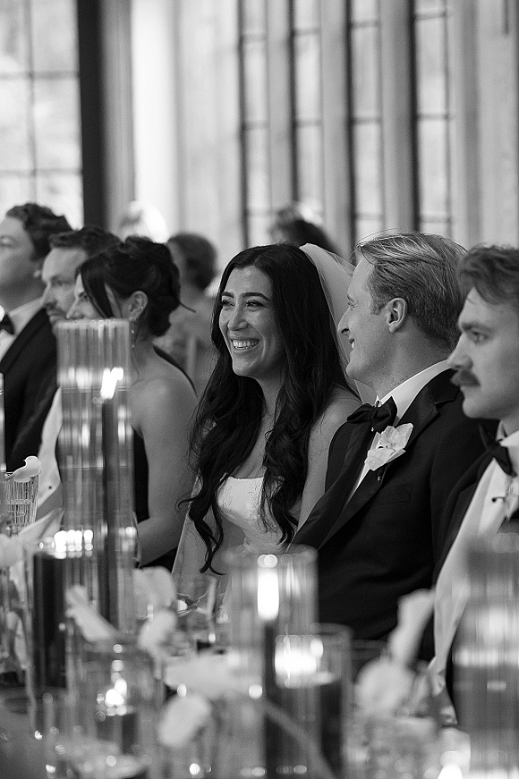 Reception moment as bride and groom at head table laugh during speeches, candlelit glass decor and guests by large windows indoors