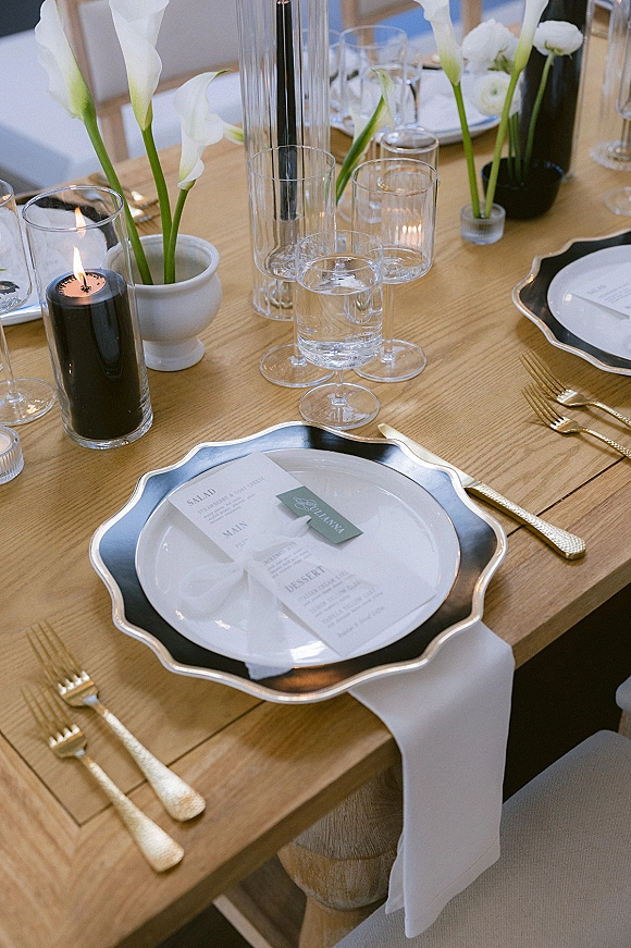 Reception tablescape with a wedding place setting featuring white calla lilies, black taper candle, gold flatware, and clear glassware on a wood table