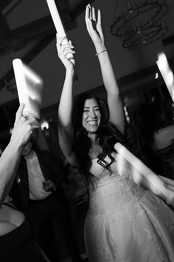 Wedding dance floor packed with guests waving wedding glow sticks as the bride in a strapless lace dress raises her hands under a chandelier