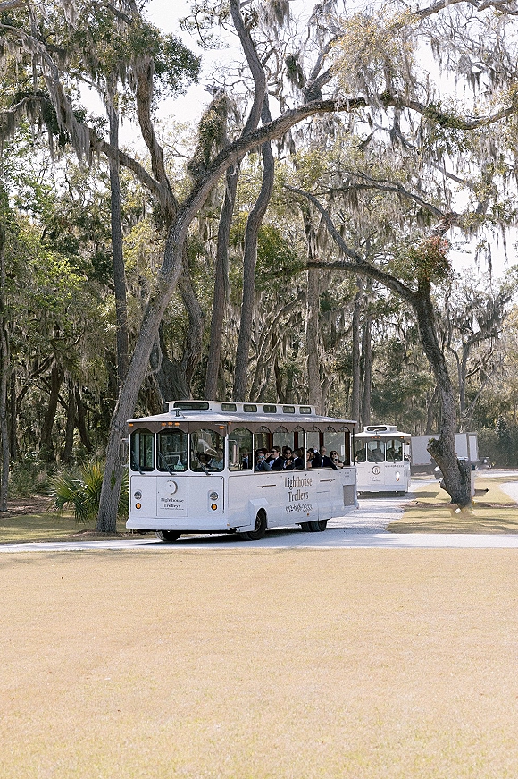 Wedding shuttle parked on a tree-lined road, a white trolley bus filled with suited guests in sunglasses beneath oak trees with hanging moss