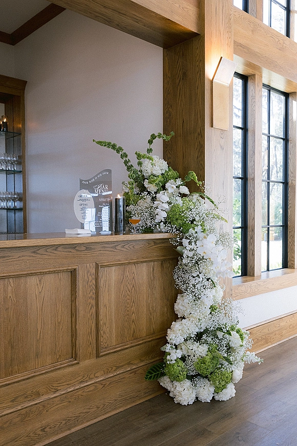Wedding bar decor with a wedding bar floral arrangement cascading hydrangeas and orchids over a wood bar, candles and menu sign by windows