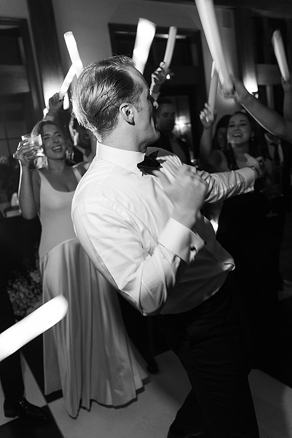 Wedding dance floor packed with wedding reception party guests waving glow sticks, one in bow tie and dress shirt holding a cocktail amid uplighting