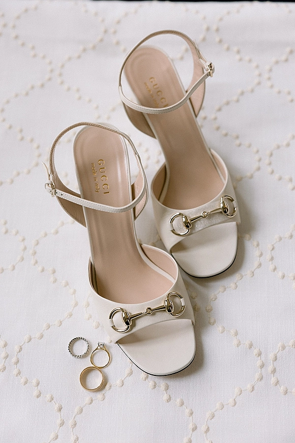 Bridal shoes in a flat lay with white bridal heels featuring gold horsebit accents, beside diamond engagement ring and gold band on quilted fabric