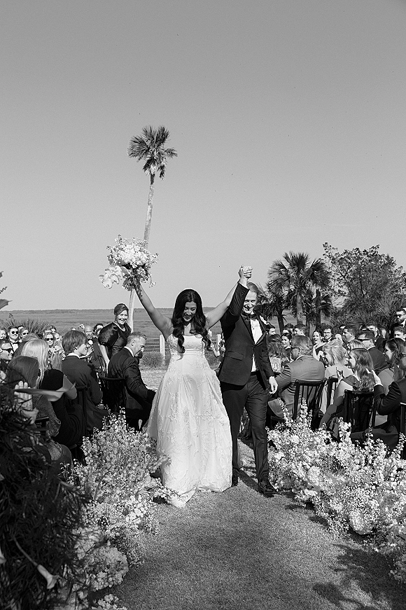 Wedding recessional as newlyweds walking down aisle, bride lifting bouquet overhead while guests cheer on a waterfront lawn with palm trees
