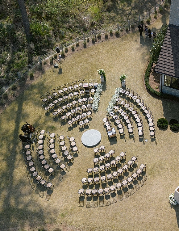Ceremony seating layout with curved chairs around a round platform, white aisle florals and pedestal arrangements on a grassy lawn with trees