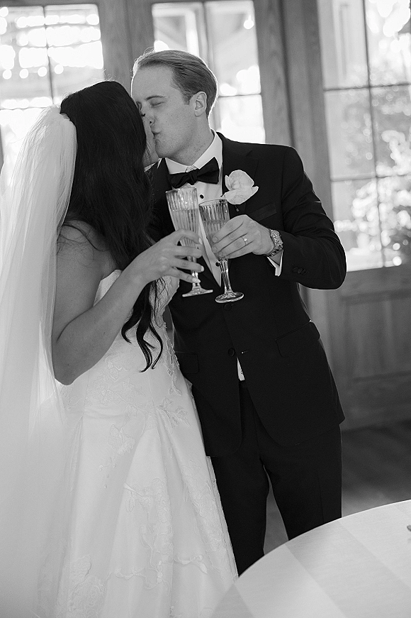 Wedding kiss portrait of newlyweds toasting champagne, bride in veil and strapless dress, groom in tuxedo by large windows at reception