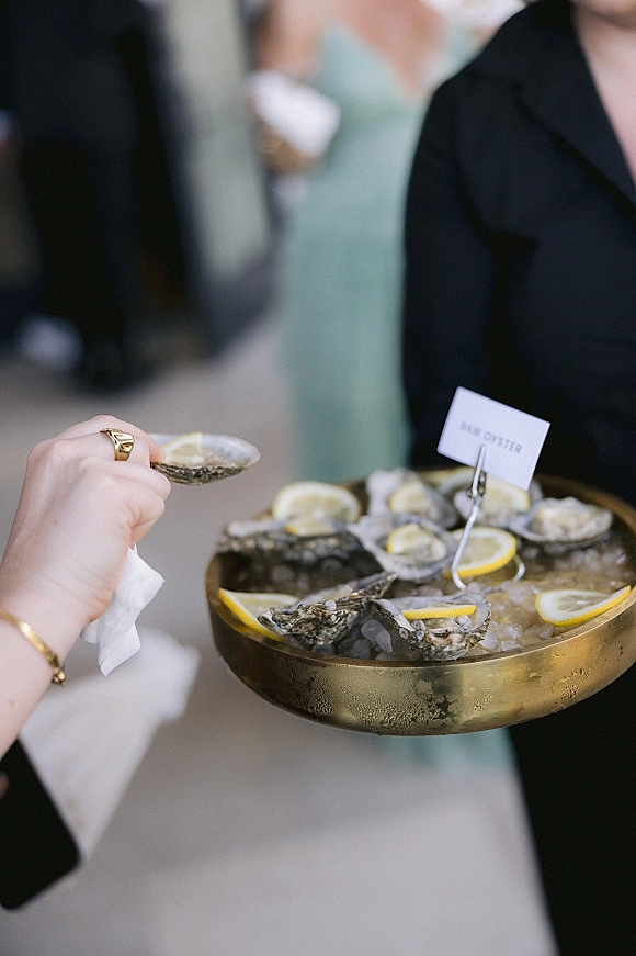 Oyster bar brass tray with shucked oysters on crushed ice and lemon wedges, gold bracelet visible amid blurred wedding guests