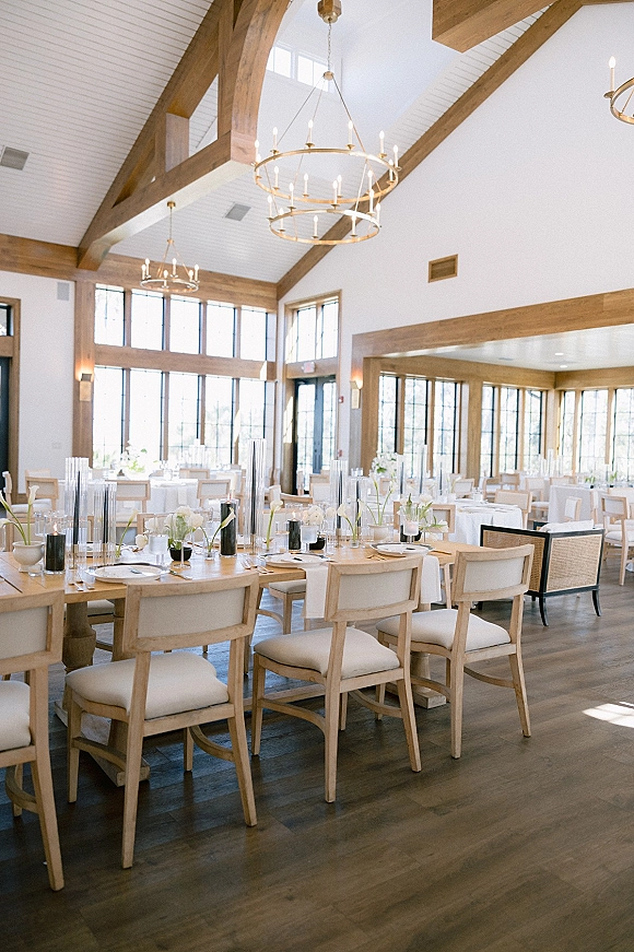 Reception tablescape with modern wedding reception decor on a long wood farmhouse table, black taper candles, white bud vases, chandeliers above in a bright windowed hall