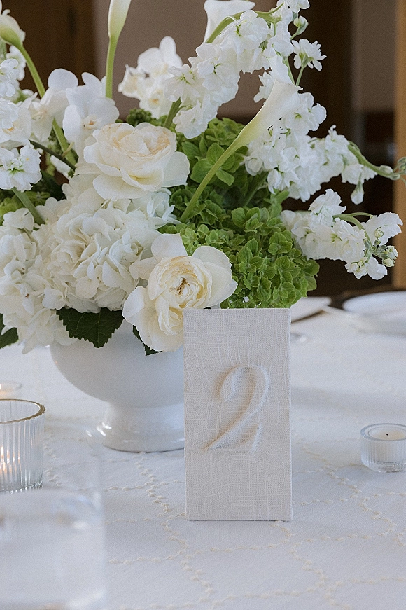 Wedding table centerpiece with white wedding centerpiece flowers in a ceramic vase beside table number 2, votive candles on a white tablecloth in a blurred reception setting