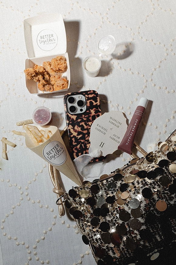 Wedding flat lay with takeout fries and fried chicken, escort card, lip gloss, tortoiseshell phone, and sequined clutch on quilted fabric