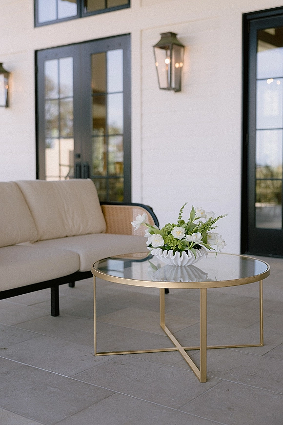 Wedding lounge decor with white floral arrangement in a ruffled bowl vase on a glass and gold coffee table beside a beige sofa on a stone patio