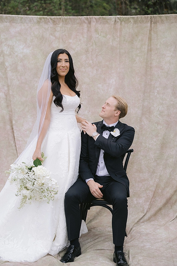 Couple portrait of bride in lace strapless gown with long veil and white bouquet, holding hands with tuxedo groom by greenery