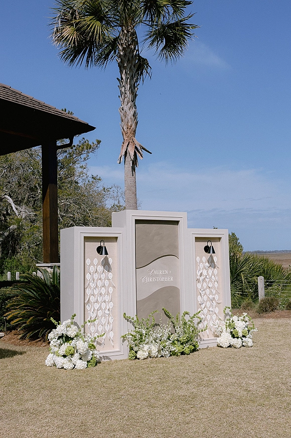 Wedding seating chart with escort card display on framed panels, flanked by white floral arrangements on a sunny lawn with palm tree backdrop