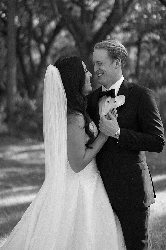 Couple portrait in a black and white wedding portrait, bride in strapless lace dress and veil holding groom’s lapel on a lawn with trees