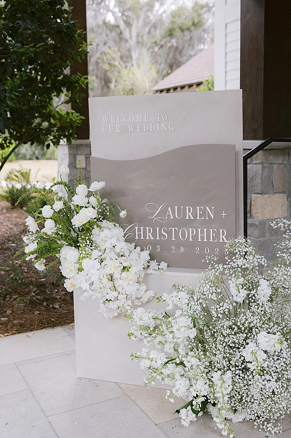 Wedding welcome sign on an acrylic arch with white florals and baby's breath, set on a porch by a stone wall and greenery