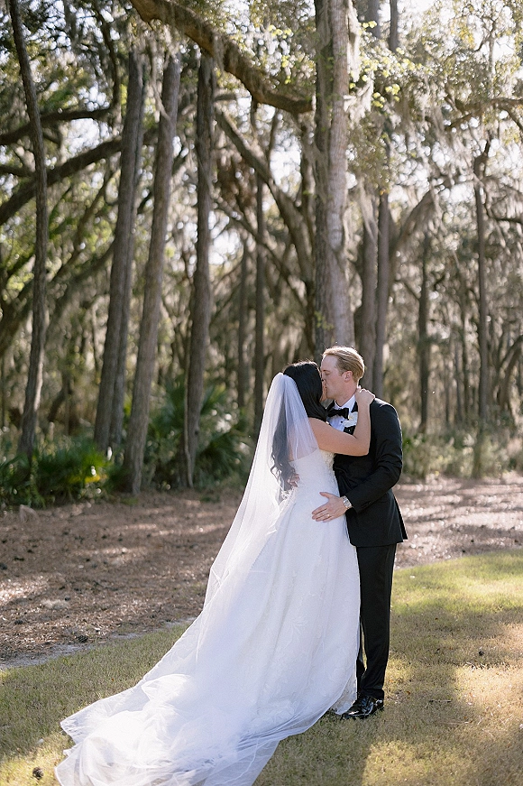 Wedding kiss portrait of bride and groom kissing, her long veil and train flowing as they embrace on a sunlit woodland path