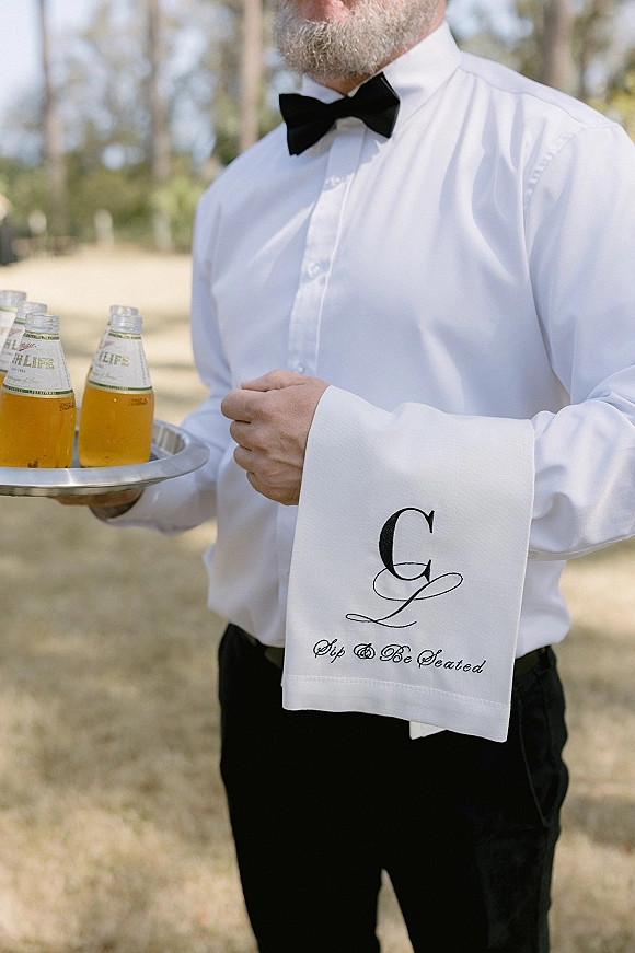 Groomsman serving drinks with a black bow tie, balancing a tray of beer bottles and monogrammed napkins on an outdoor lawn with trees