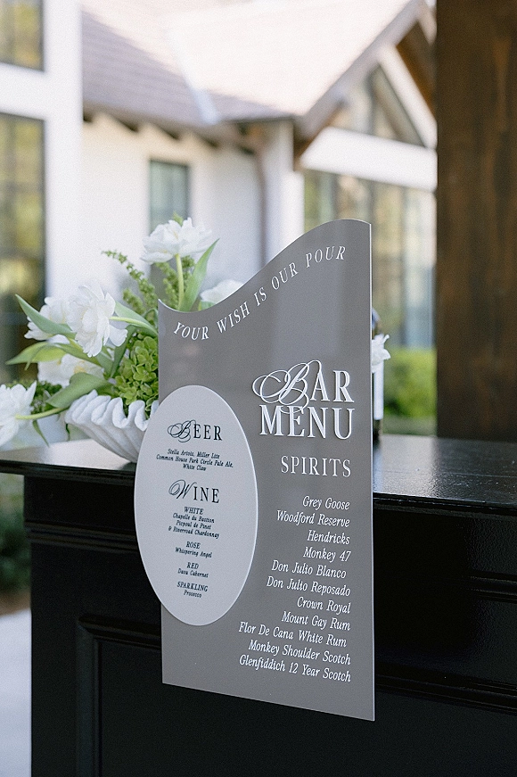 Wedding bar menu acrylic sign with signature cocktail sign styling, framed by white flowers and greenery on a tabletop outdoors