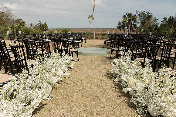 Ceremony setup for an outdoor wedding ceremony with white aisle florals and baby's breath bordering a round stage on a lawn with palms