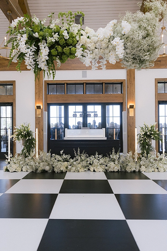Reception entrance decor with hanging floral installation of white orchids and green hydrangeas over a black-and-white checkered dance floor indoors