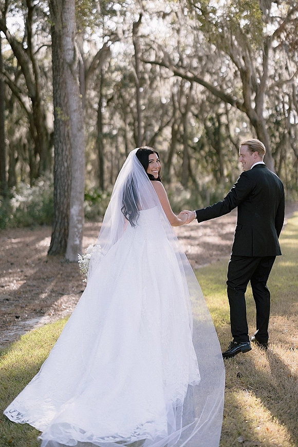Couple portrait of bride and groom holding hands on a sunlit woodland path, bride looking back with a long veil and bouquet
