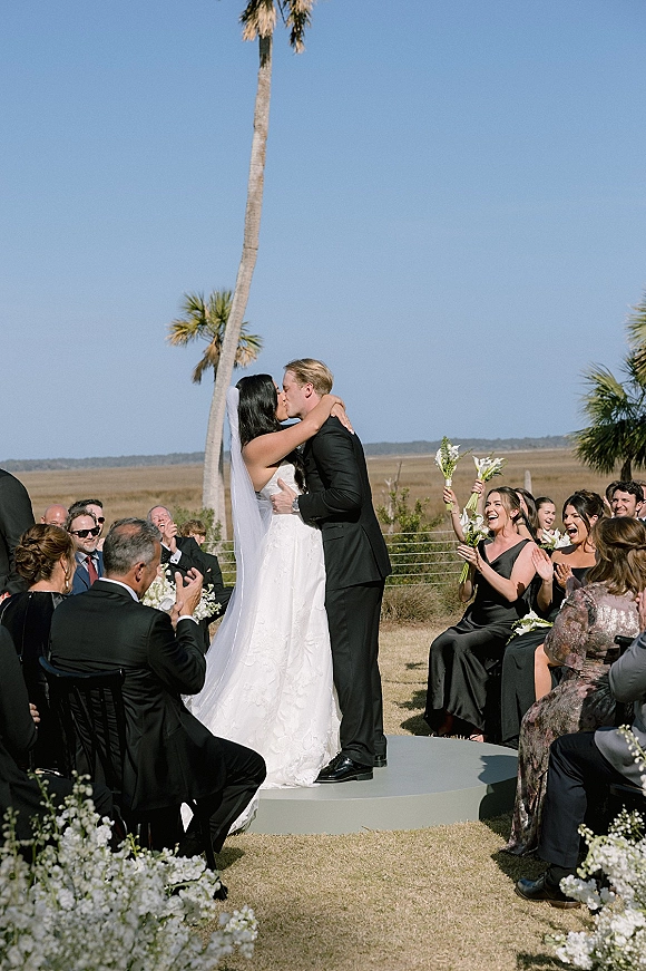 Ceremony kiss as bride and groom embrace on an outdoor wedding ceremony platform, bridesmaids cheering with white bouquets under palms