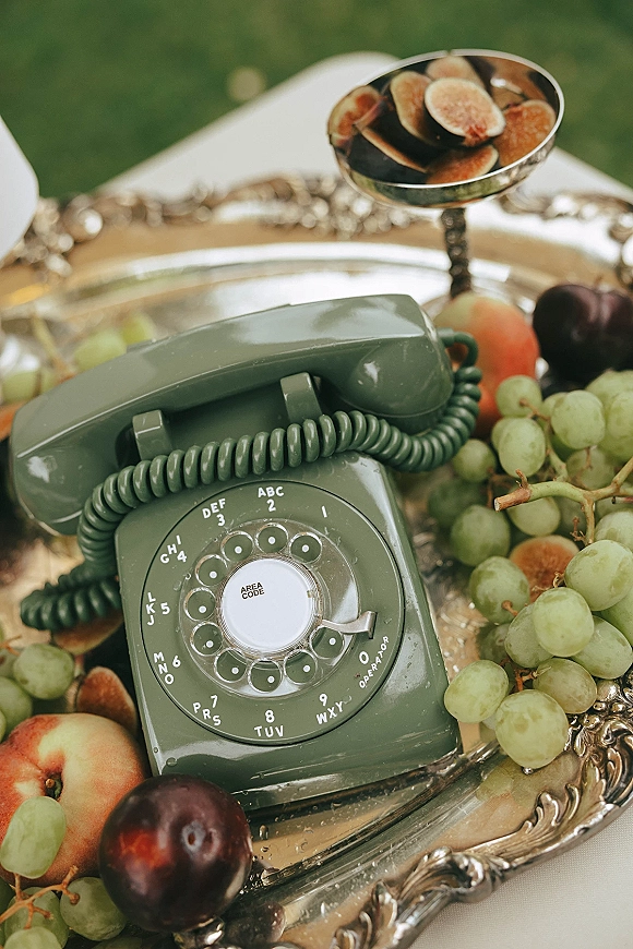 Wedding flat lay with a vintage wedding flat lay featuring a rotary phone on a silver tray with grapes, figs, and stone fruit on a lawn tablecloth