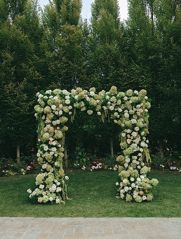 Wedding floral arch with white roses and hydrangeas, plus hanging greenery, set on a garden lawn with hedges and stone pavers