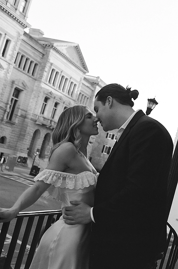 Wedding couple portrait with bride and groom touching foreheads in an off-the-shoulder lace dress by a bench on a city street near a historic facade