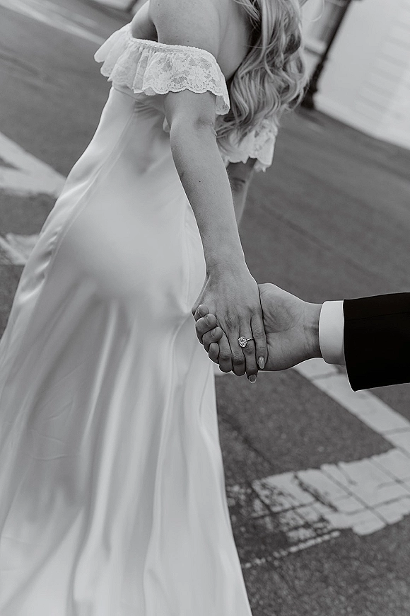 Couple holding hands with bride and groom hands showing an oval diamond engagement ring, bride’s lace sleeve, on a paved walkway by a building