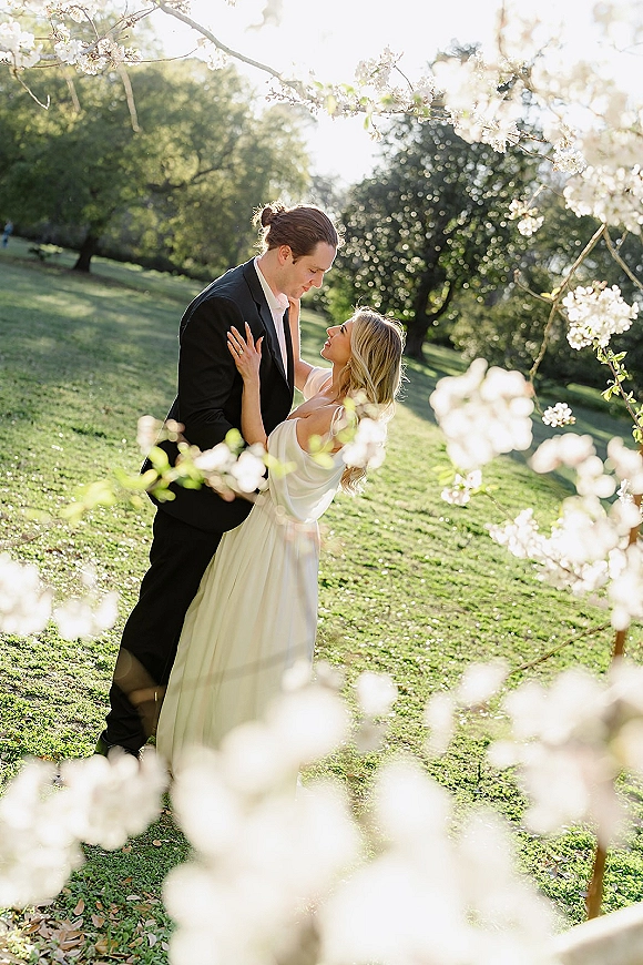 Couple portrait of bride and groom embracing under flowering tree branches, bride looking up at groom in black suit in sunlight