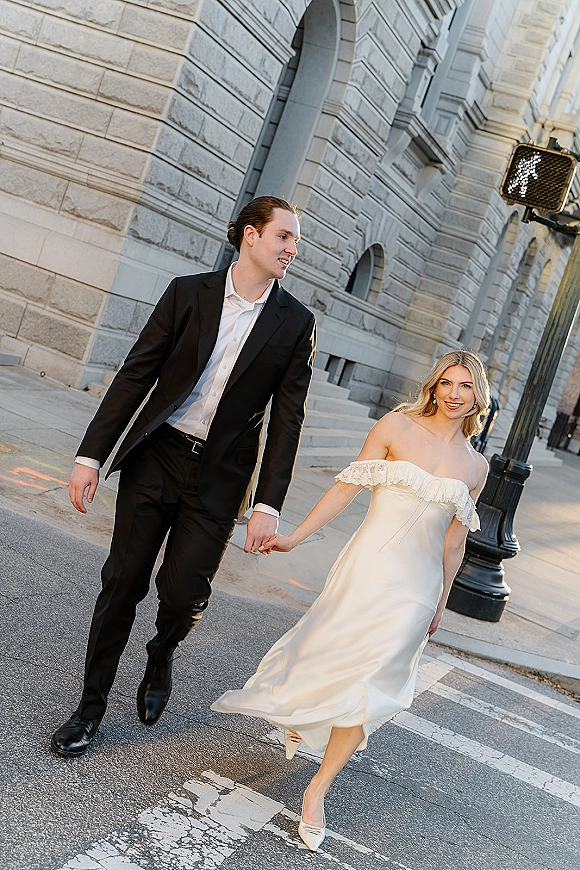Couple portrait of bride and groom walking holding hands across a city street crosswalk, her short off-the-shoulder dress and heels visible