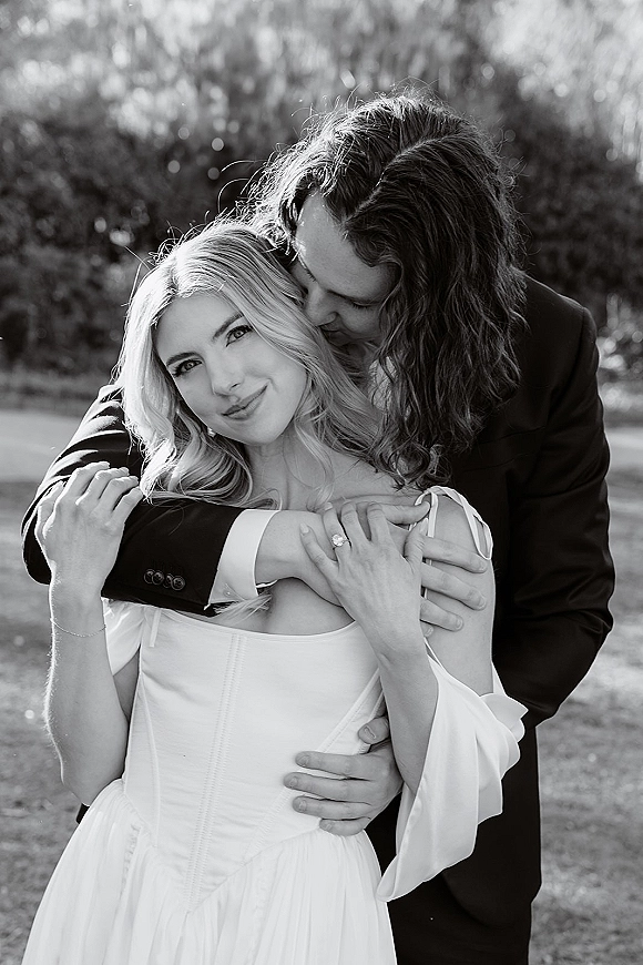 Couple portrait in black and white wedding portrait style, groom hugging bride from behind as she shows her engagement ring outdoors on a lawn