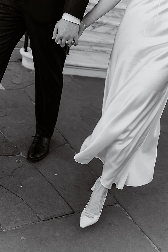 Wedding couple walking hand in hand, bride’s sleek dress hem and pointed heels beside groom’s black tux cuffs on stone steps in black and white